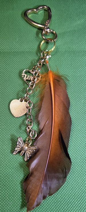 Handmade brown feather bag charm with orange feather shaft, gold-tone butterfly, textured heart, LOVE charm, chain and heart-shaped lobster clasp.